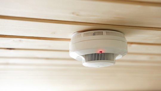 Ein weißer Rauchmelder ist an einer hellen Holzdecke montiert.