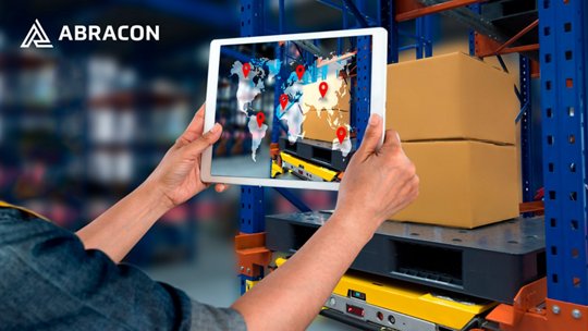A person holds a tablet in front of stacked cardboard boxes on industrial shelving in a warehouse. 