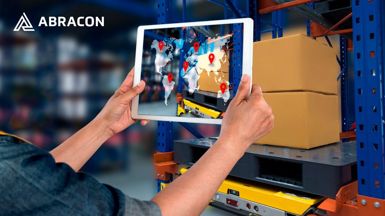 A person holds a tablet in front of stacked cardboard boxes on industrial shelving in a warehouse. 