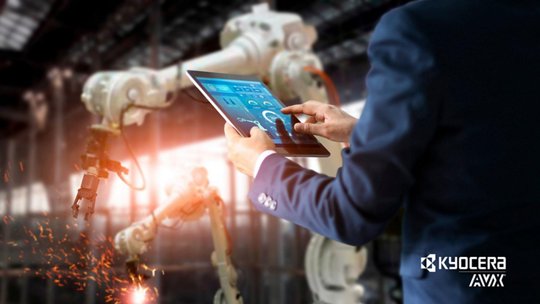 A person in a suit operates a digital tablet to control an industrial robotic arm in a modern factory environment. 