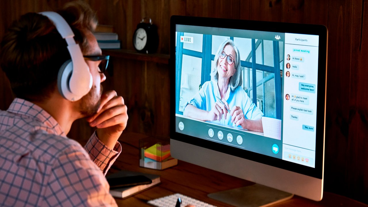 A man wearing headphones sits at a desk using a desktop computer for a video call. 