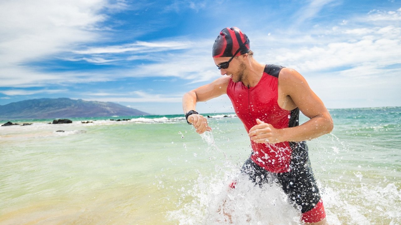 A male athlete wearing a red and black sports outfit runs through shallow ocean waves on a sunny day