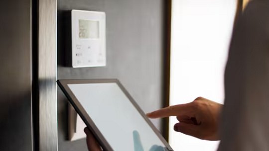 A person interacts with a digital tablet in front of a wall-mounted smart thermostat.