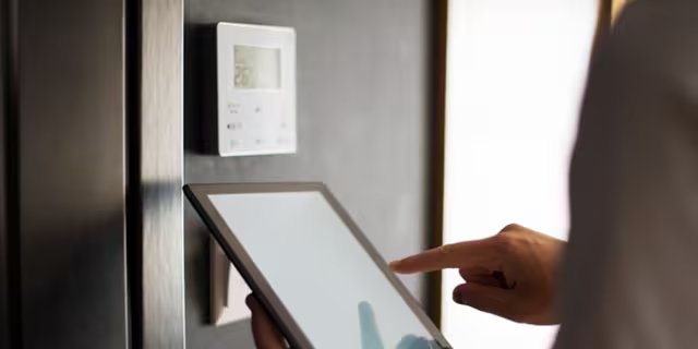A person interacts with a digital tablet in front of a wall-mounted smart thermostat.