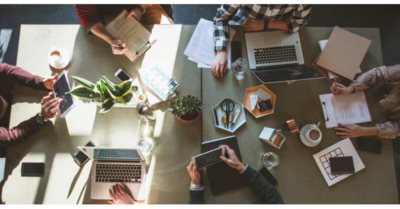 A group of people are gathered around a large table, working together using laptops, tablets, and notepads.