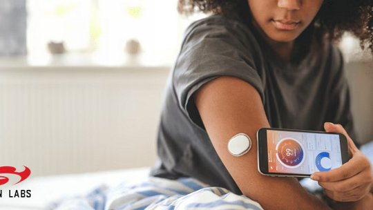 A young woman interacts with a health monitoring device attached to her arm while viewing data on a smartphone app. 