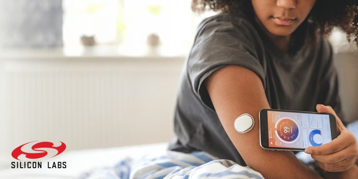 A young woman interacts with a health monitoring device attached to her arm while viewing data on a smartphone app. 