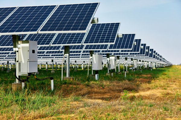 Ground-mounted solar panels arranged in long rows across a grassy field, with gray inverter boxes and electrical conduits installed beneath each panel structure, extending into the distance under a clear sky.