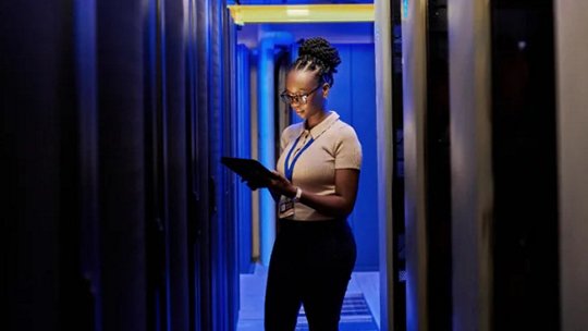 A person stands in a corridor between server racks, holding a tablet and wearing a lanyard. The setting is a high-tech data center with blue ambient lighting and visible server equipment.