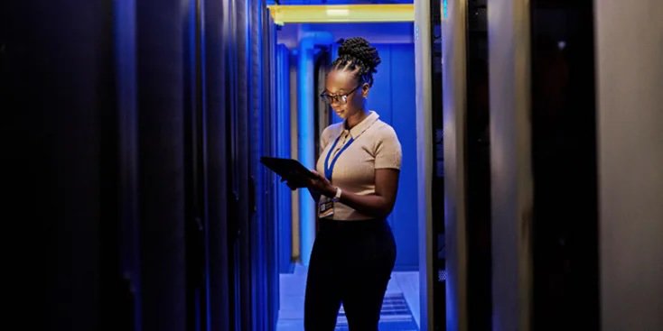 A person stands in a corridor between server racks, holding a tablet and wearing a lanyard. The setting is a high-tech data center with blue ambient lighting and visible server equipment.