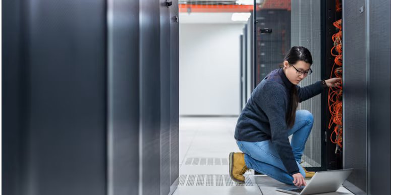 A person is kneeling on the floor of a data center, working on a laptop while connecting or troubleshooting network cables in a server rack. The environment is clean and high-tech, with rows of server cabinets and visible orange cabling.