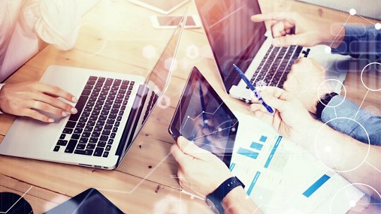 A group of people work together at a wooden table, using laptops and a digital tablet. 