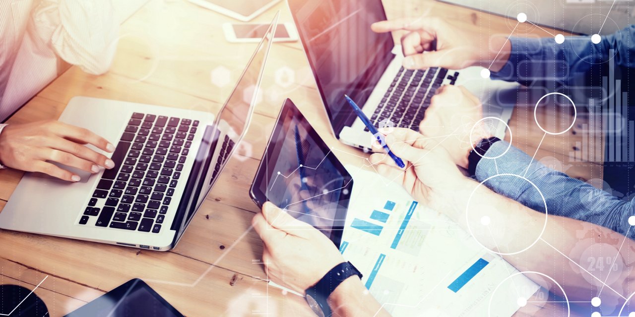 A group of people work together at a wooden table, using laptops and a digital tablet. 