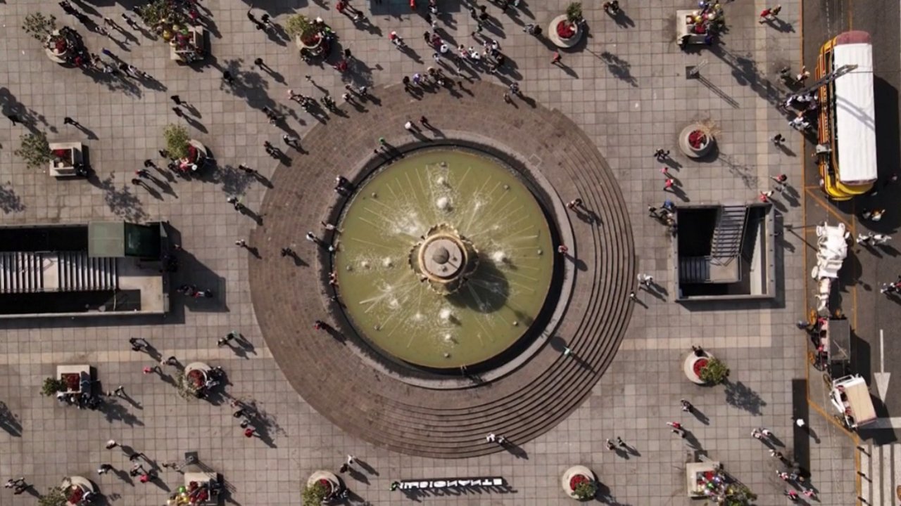This overhead image captures a large circular fountain at the center of a bustling urban plaza. 