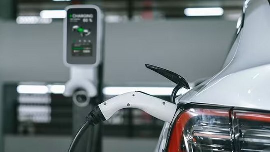 A white electric vehicle is shown plugged into a charging station in an indoor facility. The focus is on the charging cable connected to the car's rear, with the charging unit and display visible in the background.