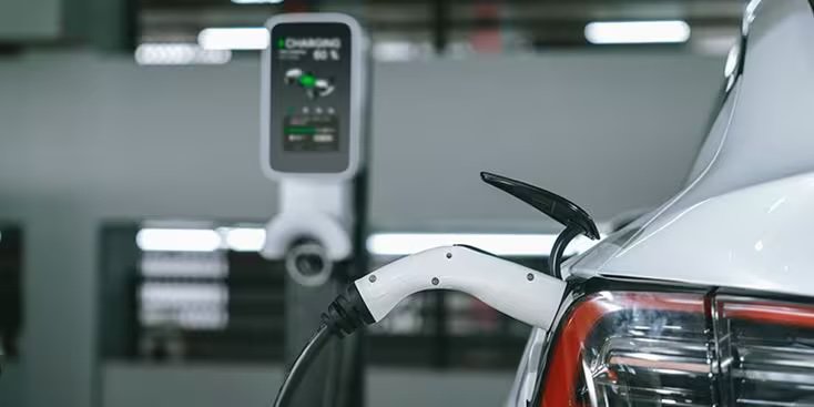 A white electric vehicle is shown plugged into a charging station in an indoor facility. The focus is on the charging cable connected to the car's rear, with the charging unit and display visible in the background.