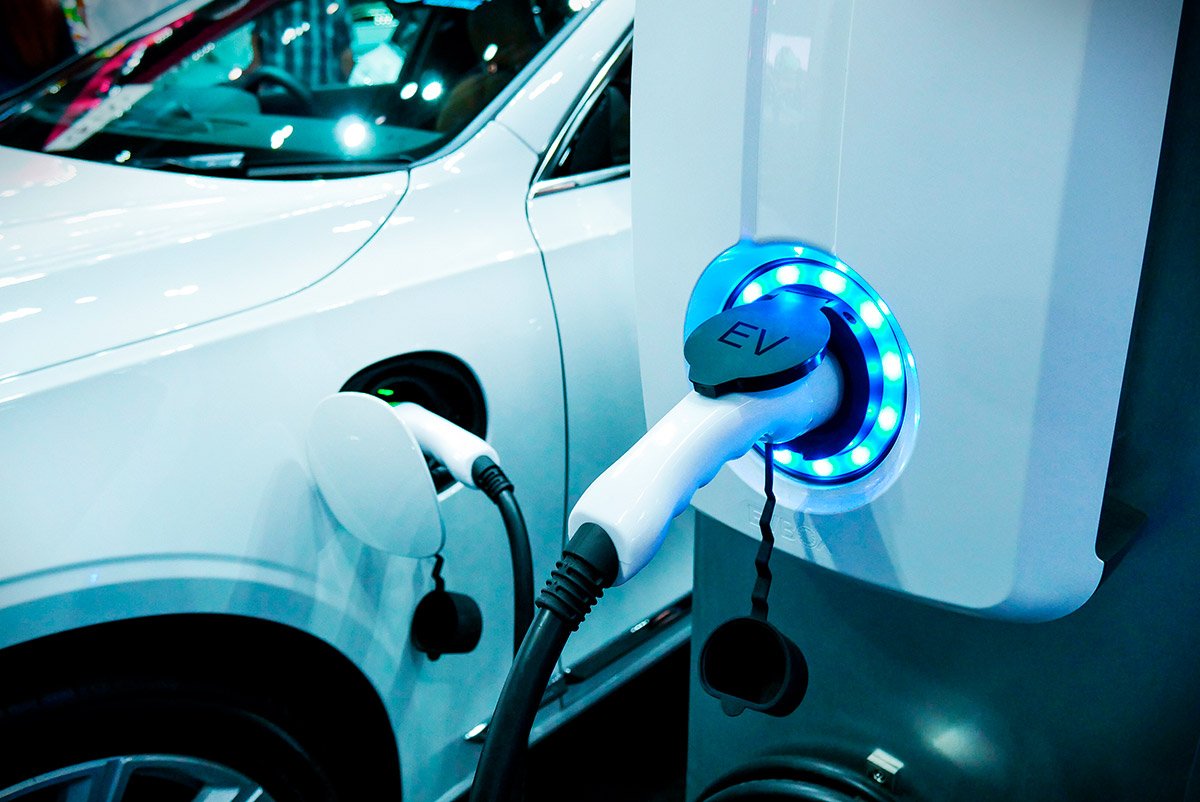 A white electric vehicle is connected to a charging station via a plugged-in charging cable. 