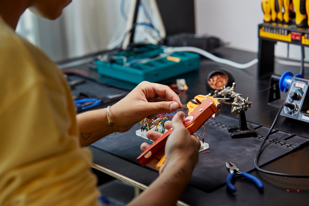 Primo piano di una persona che utilizza una spelafili per preparare piccoli cavi elettrici su un banco da lavoro con strumenti per l’elettronica, attrezzature per la saldatura e componenti, suggerendo la realizzazione pratica di un prototipo o l’assemblaggio di un circuito.