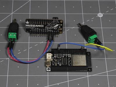 ESP32 development board connected by jumper wires to a Feather DALI interface board and terminal connectors, arranged on a cutting mat.