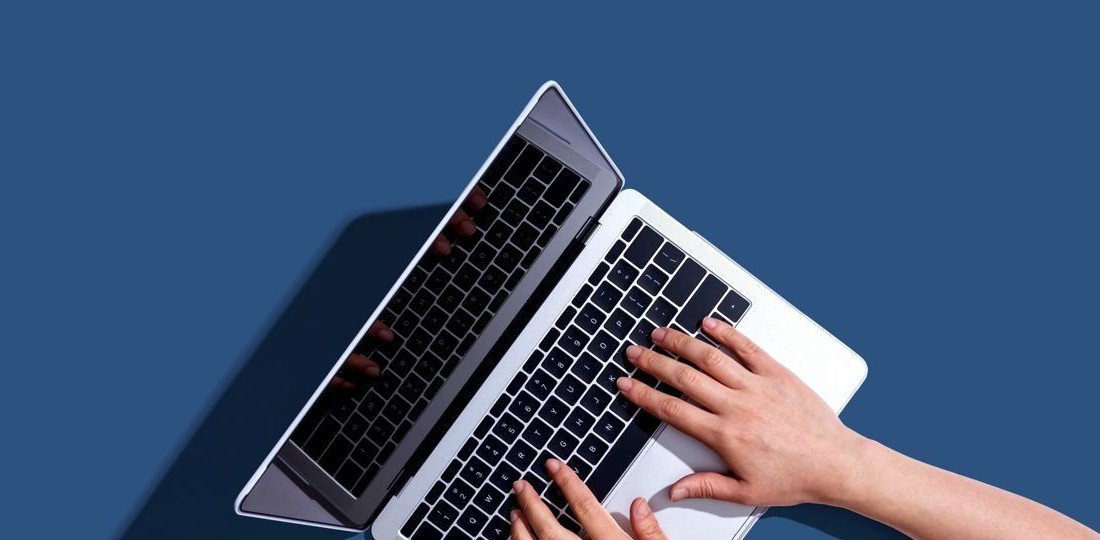A pair of hands is seen typing on a sleek laptop keyboard against a solid blue background.