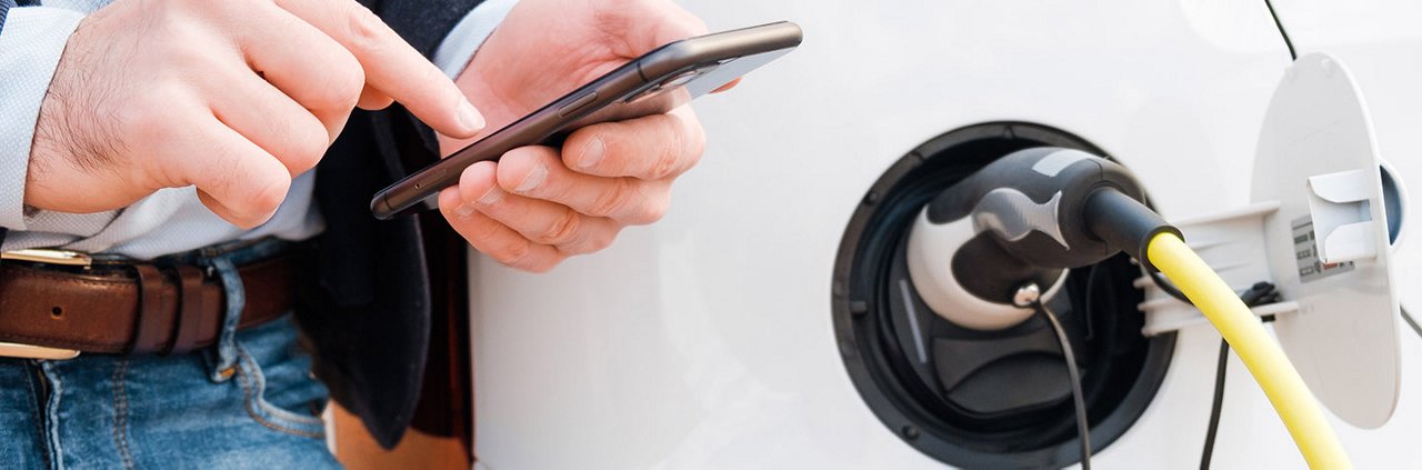 A close-up of a man holding a smartphone while standing near an electric car charging station. 