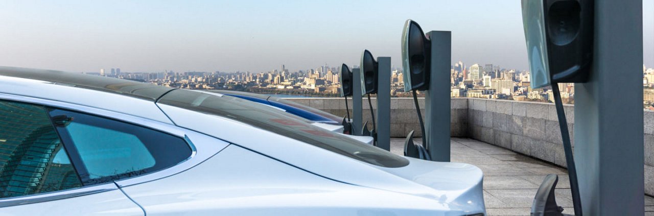 A row of electric cars parked at a rooftop charging station with a city skyline in the background. 