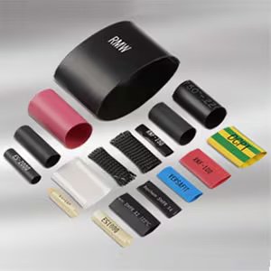 A variety of heat shrink tubing pieces are arranged on a neutral background. The tubes come in different colors such as black, red, yellow, green, blue, and white, with some featuring visible text like 'RMW', '407-22', 'RNF-100', and 'ESTI 105°C'.