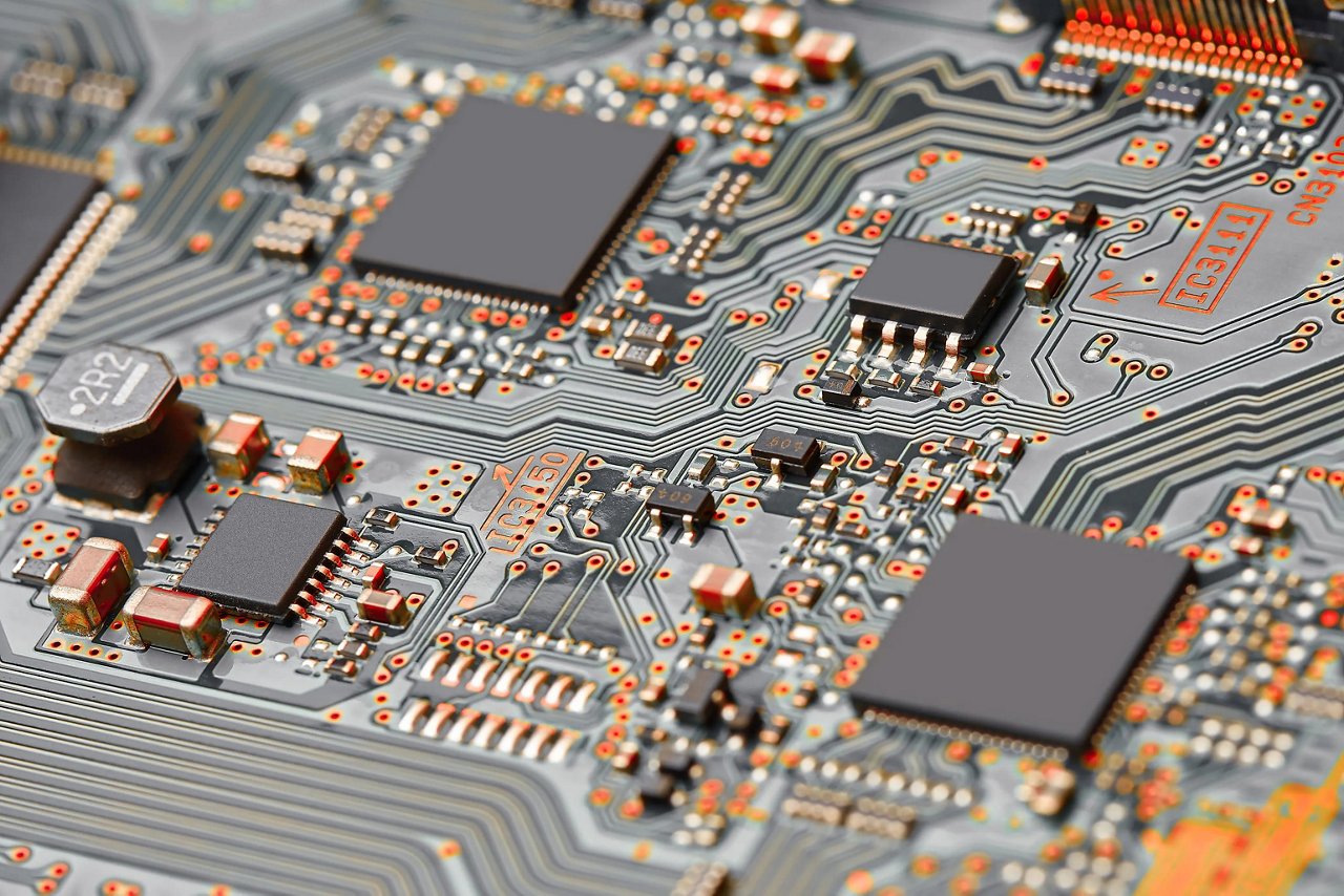 Close-up view of a printed circuit board with mounted integrated circuits, resistors, and capacitors, showing fine metal traces and solder points connecting electronic components across the board surface.