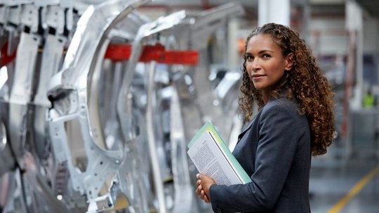 Person standing in an automotive manufacturing plant, holding documents beside assembled car body frames on a production line, with metal chassis components and industrial equipment extending into the background under factory lighting.
