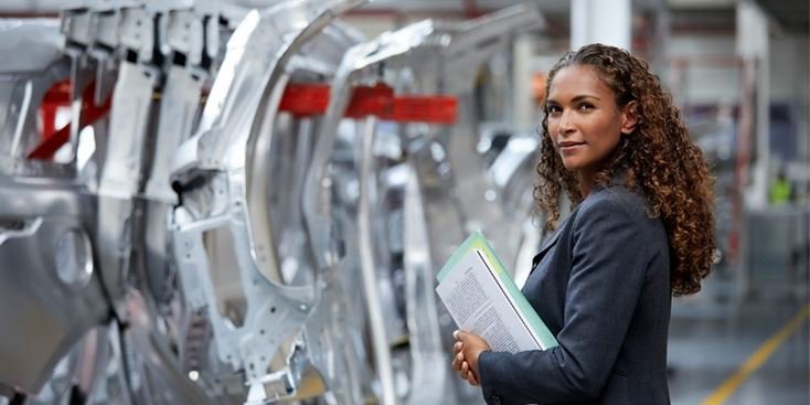 Persona in piedi in un impianto di produzione automobilistica, con documenti in mano accanto a scocche di automobili assemblate su una linea di produzione, con componenti di telaio in metallo e attrezzature industriali che si estendono sullo sfondo sotto l'illuminazione della fabbrica.