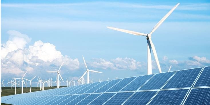 Pannelli solari in primo piano con più turbine eoliche su un paesaggio pianeggiante sotto un cielo blu con nuvole sparse, che illustrano la generazione di energia rinnovabile da fonti solari ed eoliche.