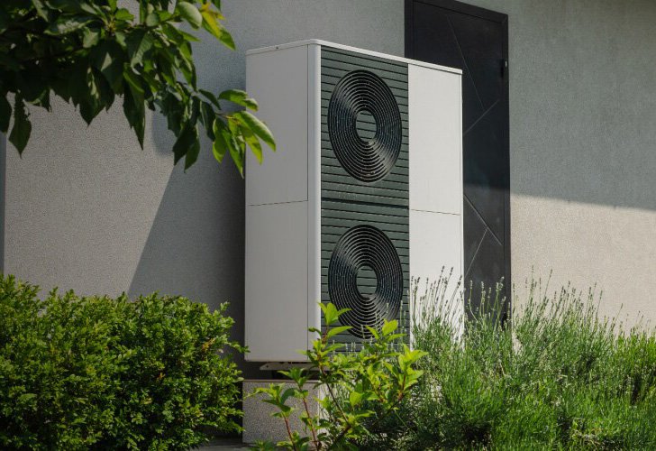 An outdoor heat pump unit with two large circular fans is mounted on the exterior wall of a modern building. 