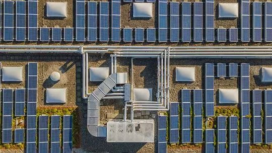 An array of solar panels installed on a flat rooftop, captured from above. Prominent industrial pipes and ventilation systems run through the center, surrounded by evenly spaced solar modules.