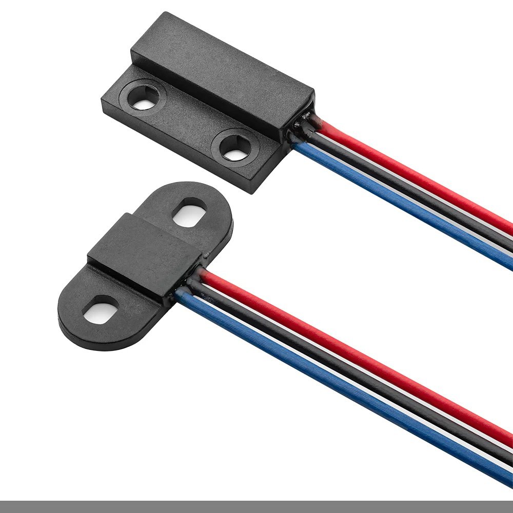 A close-up image of a magnetic reed switch sensor, featuring two black plastic components with mounting holes and attached red, blue, and black wires.