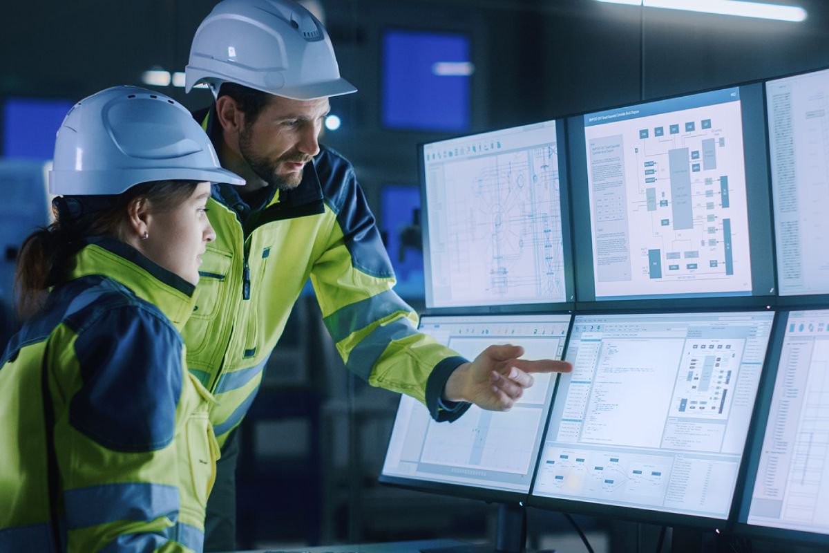 Two engineers in high-visibility jackets and helmets review complex data displayed on several large monitors in a control room. 