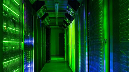 A brightly lit corridor in a modern data center features rows of server racks glowing with green and blue LED lights. The environment is highly controlled, with visible ventilation and cable management overhead.