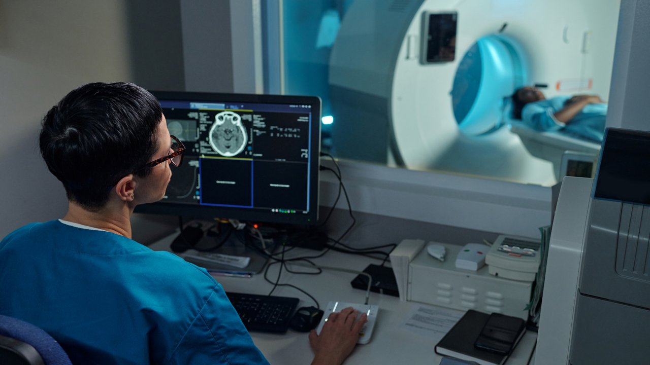 A medical technician observes brain scan images on a computer screen while a patient undergoes an MRI scan in a clinical setting. 