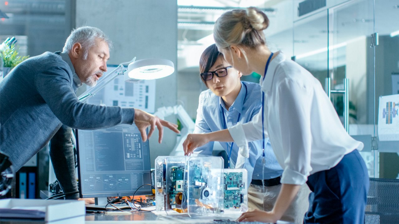A group of professionals gathers around a desk, closely examining and discussing a transparent computer hardware prototype. 