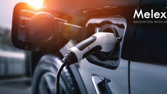 Un véhicule électrique est montré en train de charger, avec un gros plan sur le port de recharge et le câble. La scène semble se dérouler en extérieur pendant la journée, avec une lumière chaude du soleil. Le logo Melexis et le slogan "Innovation with Heart" sont clairement visibles dans le coin supérieur droit.