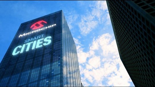 A modern skyscraper displays the text 'Microchip Smart Cities' prominently on its glass facade. The building is set against a bright blue sky with scattered clouds, emphasizing urban innovation. 