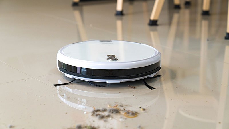 A sleek white robotic vacuum cleaner is actively cleaning a dusty tile floor in a modern indoor setting. 