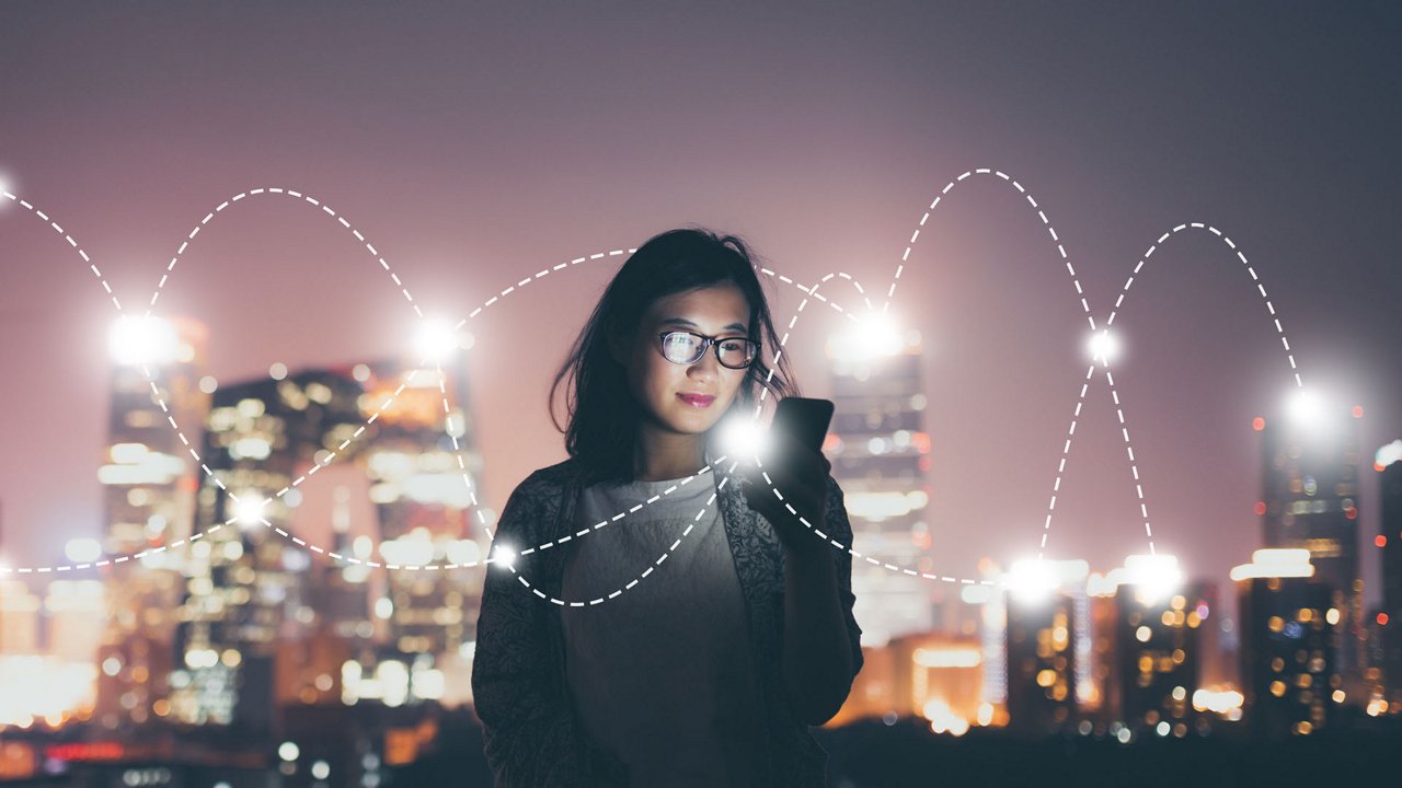 A person holds a smartphone while standing against a vibrant cityscape at night.