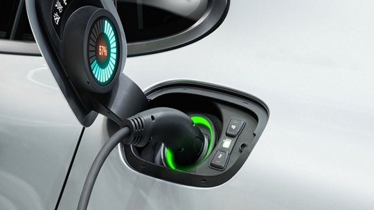 Close-up of an electric vehicle charging port with a connected cable. The charging interface displays a glowing green light and a visible numeric indicator reading '57%'.