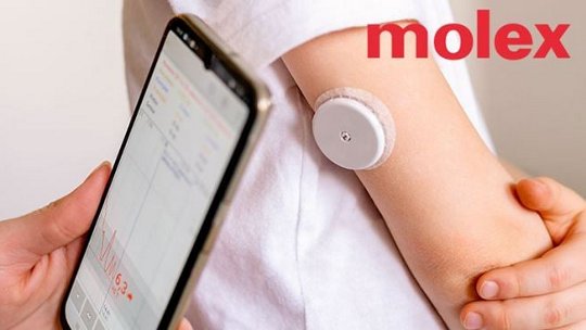 A wearable medical sensor is attached to a person's arm, paired with a smartphone displaying health data. The Molex logo is clearly visible in the image. The phone screen shows a graph and numeric data, including '6.3'. The setting suggests a health monitoring activity.
