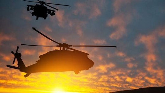Two helicopters are silhouetted against a vibrant sunset sky, creating a dramatic and dynamic scene. 