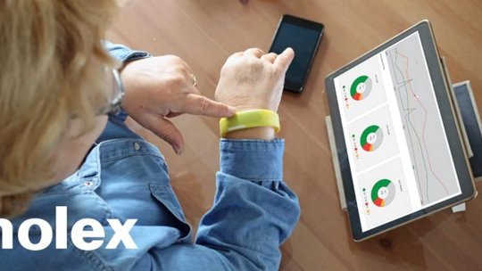 An older woman interacts with a yellow wearable device on her wrist while seated at a wooden table