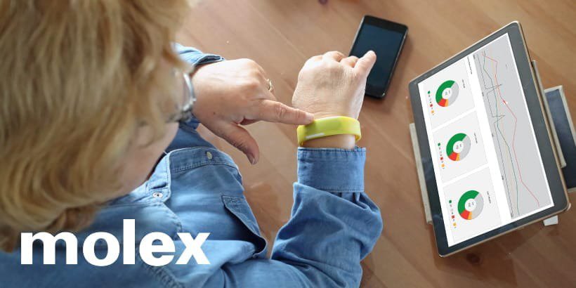 一位年长女性坐在一张木桌旁，与她手腕上的黄色可穿戴设备互动。