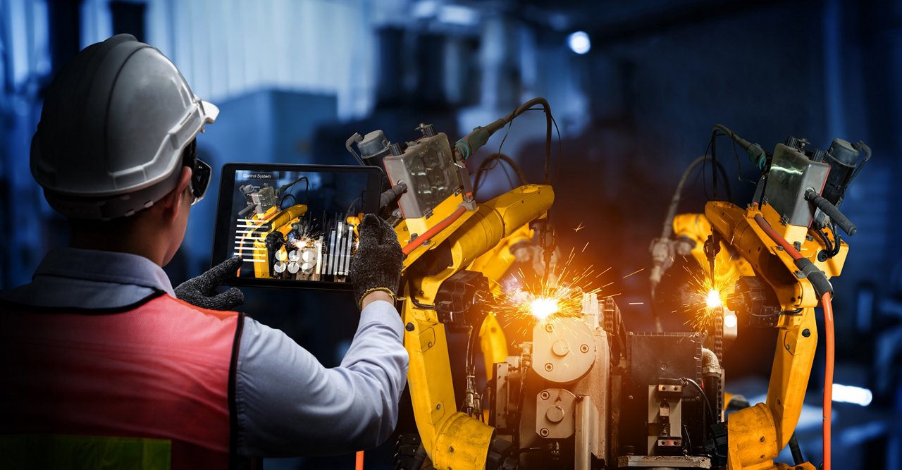 A factory engineer wearing a safety helmet and vest uses a tablet to monitor robotic arm