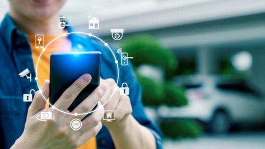 A close-up of a person holding a smartphone, interacting with smart home icons displayed digitally around the device.