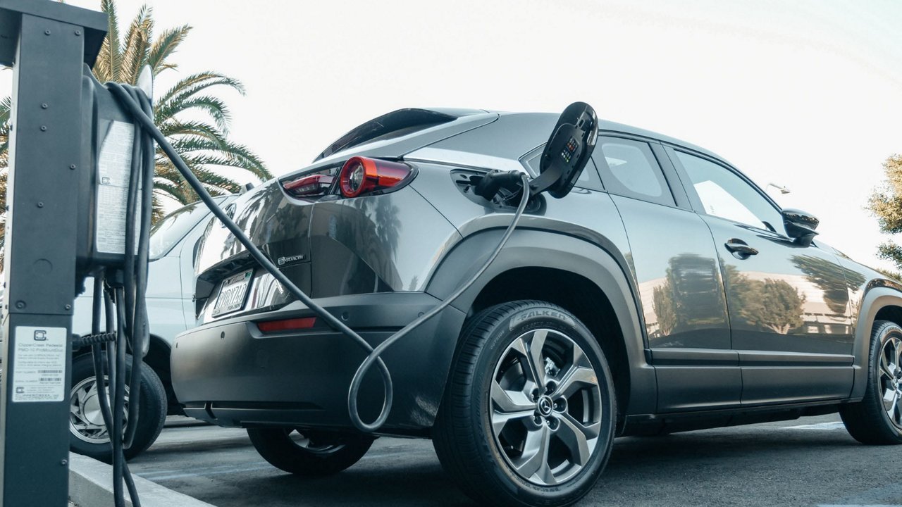A modern electric SUV is connected to a charging station in an outdoor parking area. The vehicle is gray with sleek design features, and the charging cable is visibly plugged into its port. 